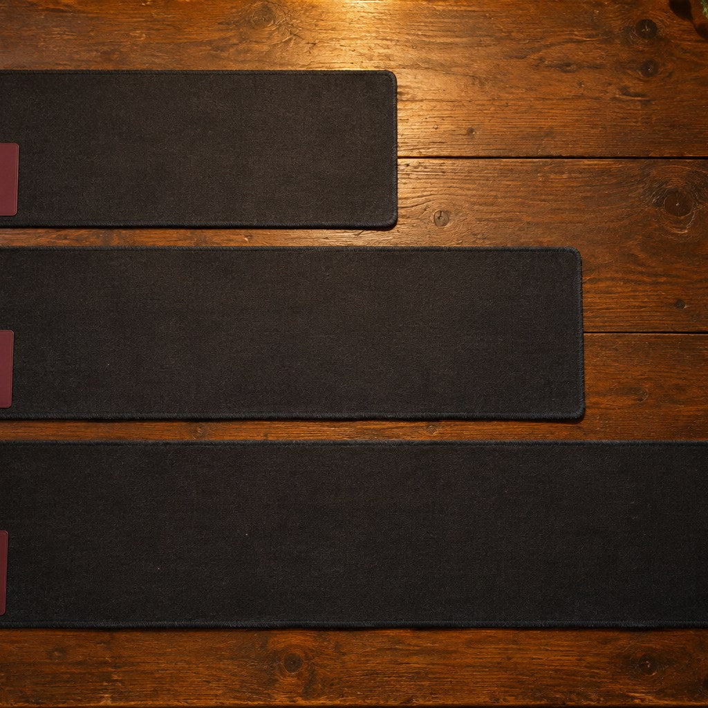Three plain dark playmats of increasing length lined up side by side on a wooden table, each with one card in its corner.