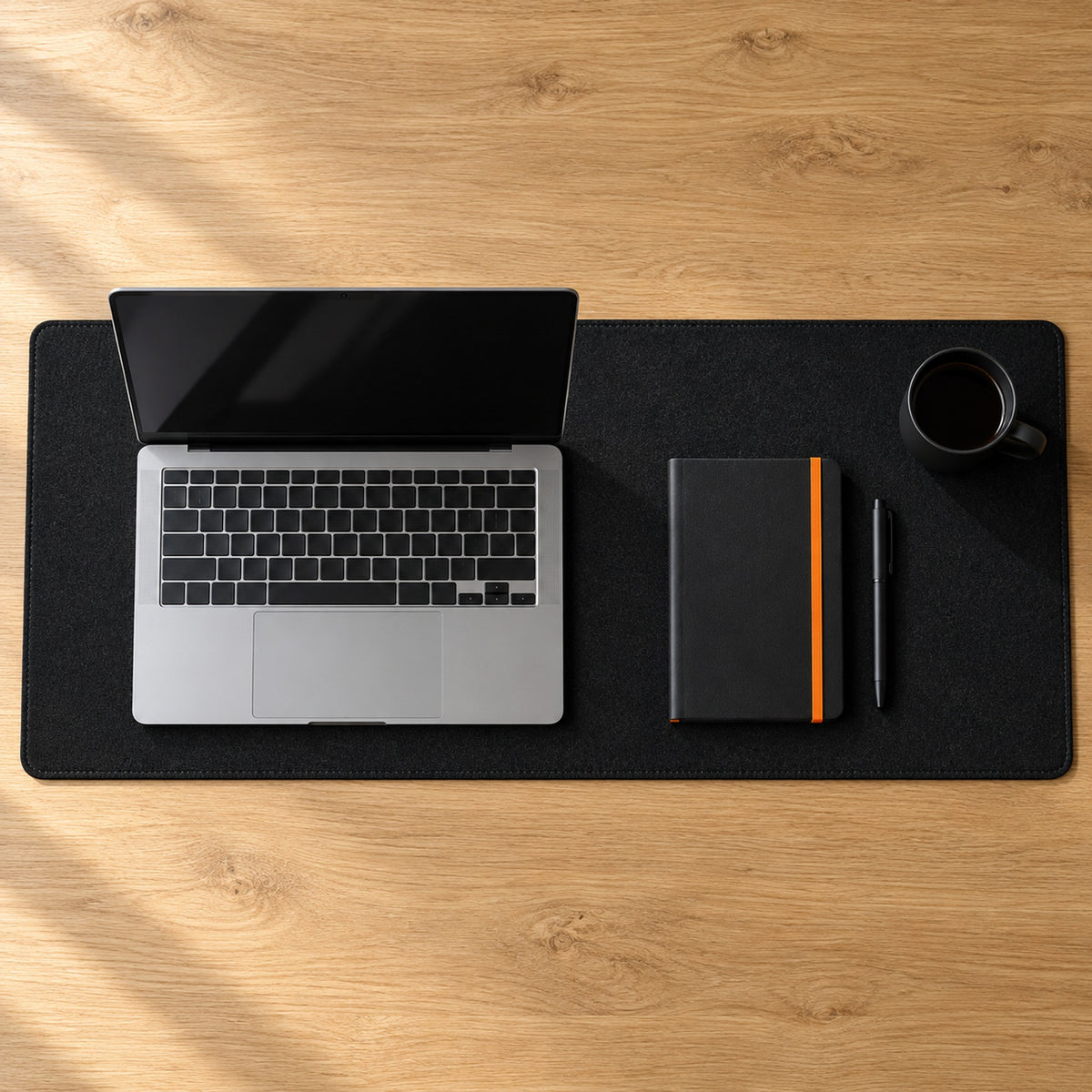 Black felt desk pad from above on a light oak desk with a laptop, black notebook, pen and cup
