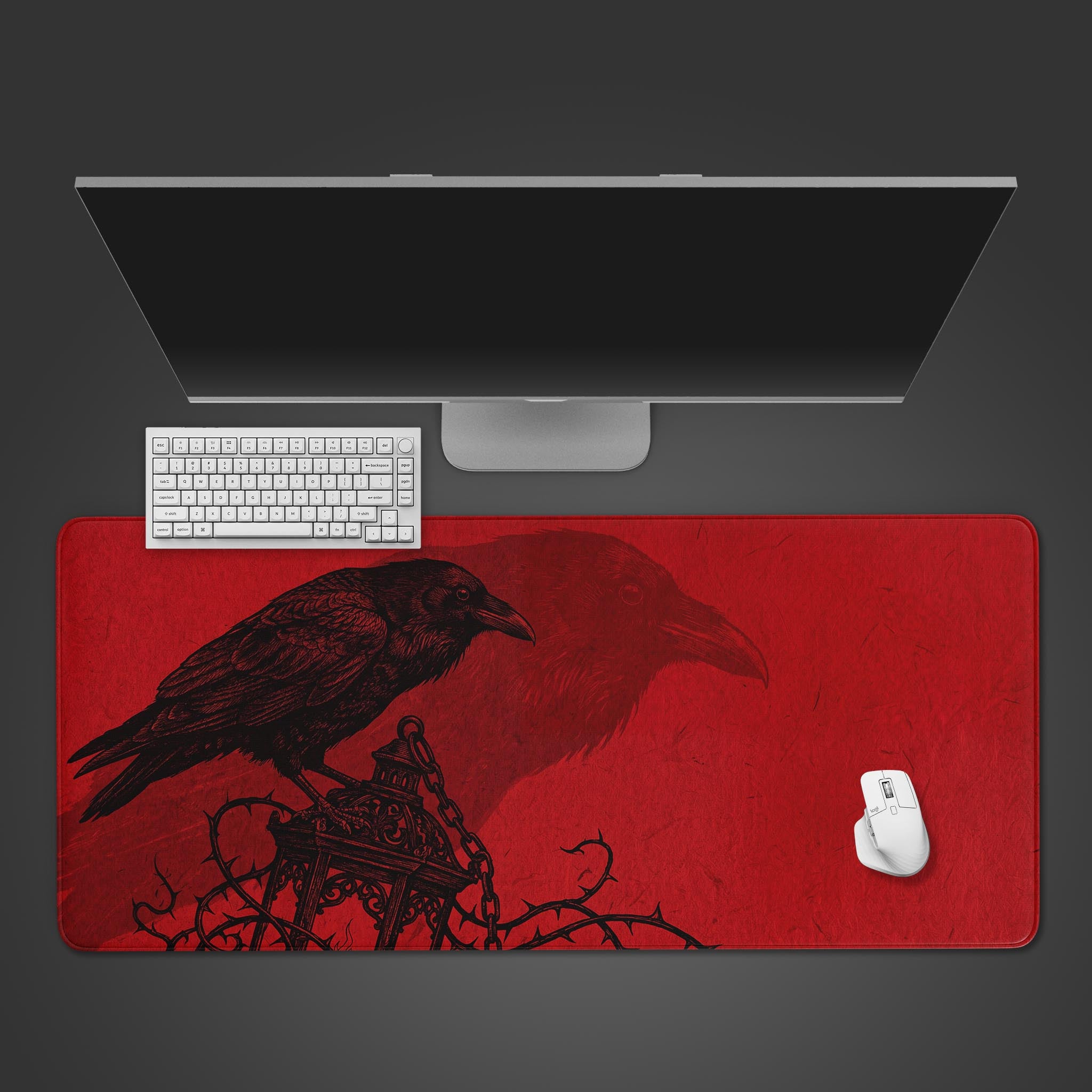 Top-down view of the Crimson Raven gaming desk mat, showing the gothic raven design alongside a white keyboard, mouse, and a large computer monitor on a dark grey surface. - Epic Design Pads & Mousepads