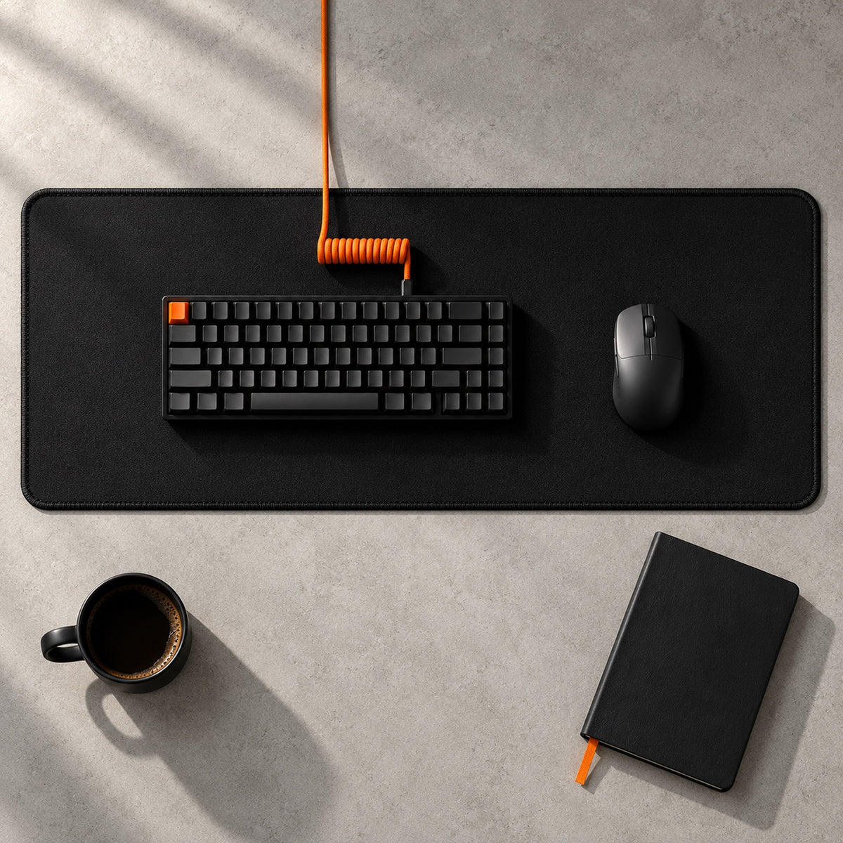 Pure Black Desk Pad from above on a concrete desk, with a black keyboard, orange coiled cable and black mouse; black stitched edge all round