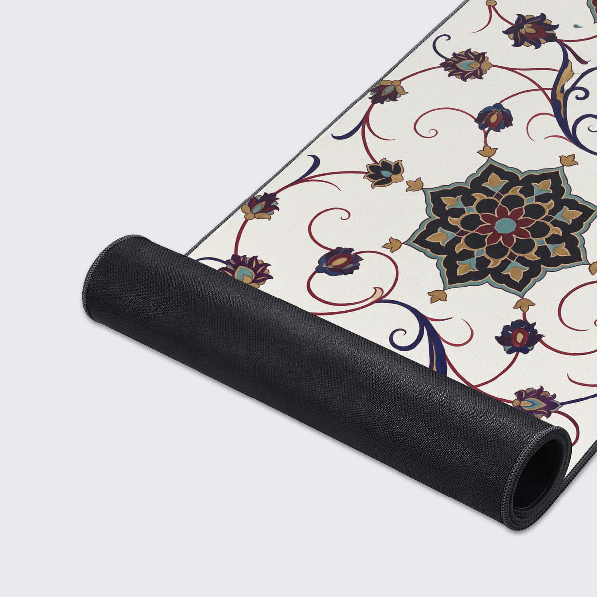 Desk mat partly rolled up, showing its black non-slip rubber underside and ivory art with a dark floral medallion and vines