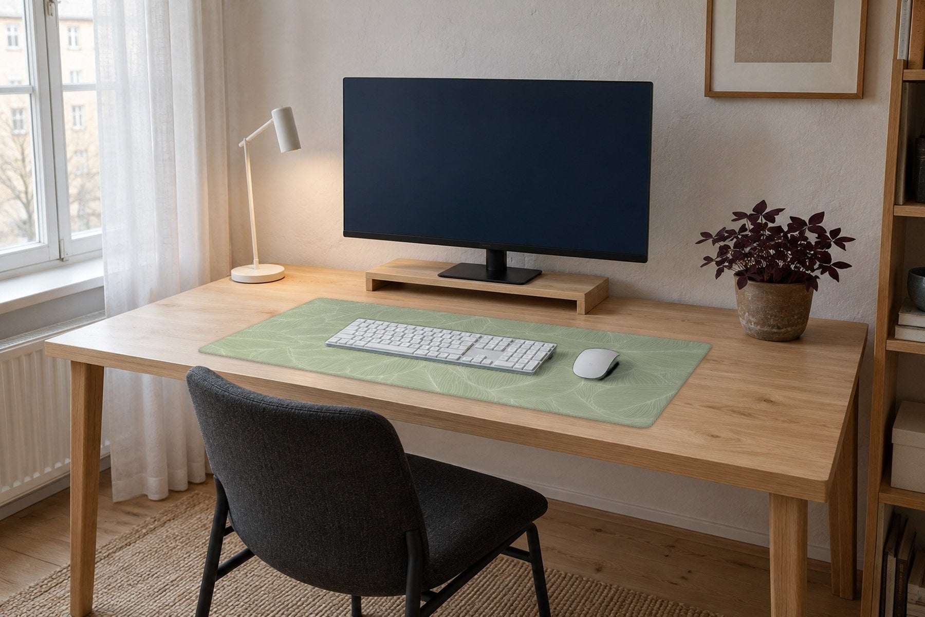 A bright home office with a pale oak desk by a window, a sage green desk mat with white leaf line art under a white keyboard and mouse, and a plant and lamp beside the mat.