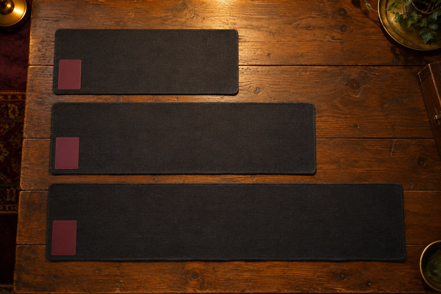 Three plain dark playmats of increasing length lined up side by side on a wooden table, each with one card in its corner.