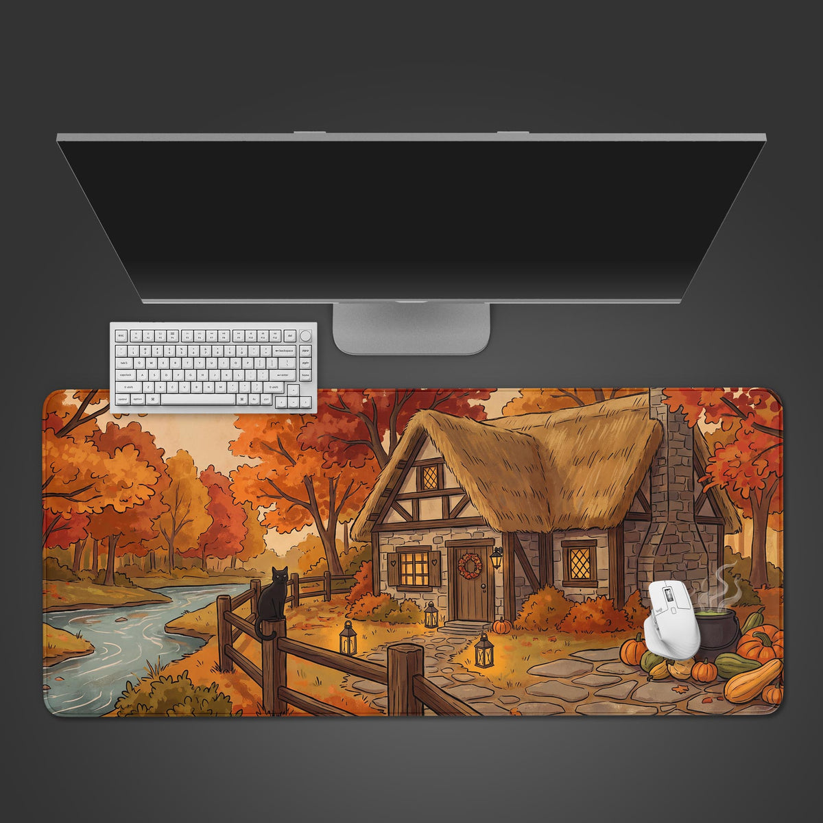 A large desk mat displaying a cozy autumn cottage scene with a black cat on a fence, pumpkins, and a cauldron by a stream.