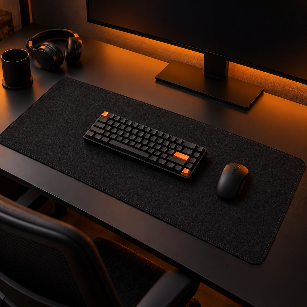 Black felt desk pad on a dark desk lit orange behind the monitor, with keyboard, mouse, cup and headphones