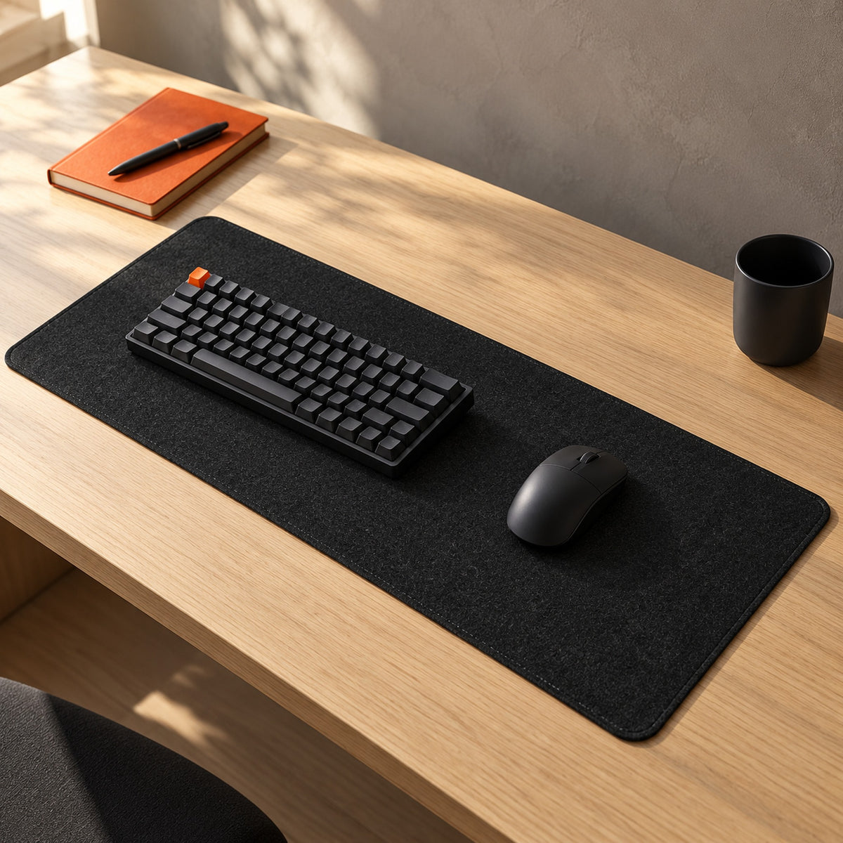 Black felt desk pad on a light oak desk in daylight, with keyboard, mouse, orange notebook and black cup