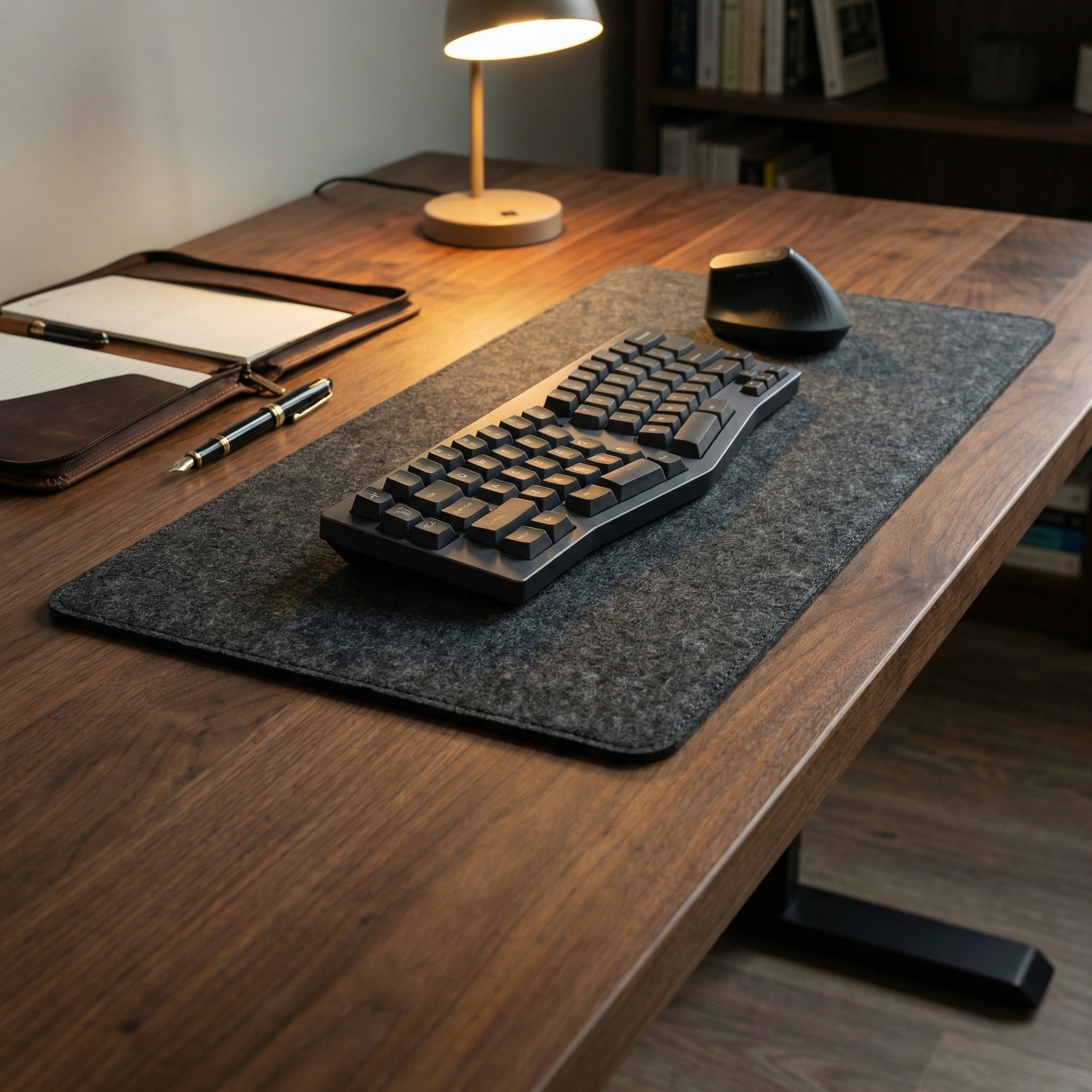 Wooden desk with keyboard, mouse, and lamp on a large felt desk mat in a home office setting