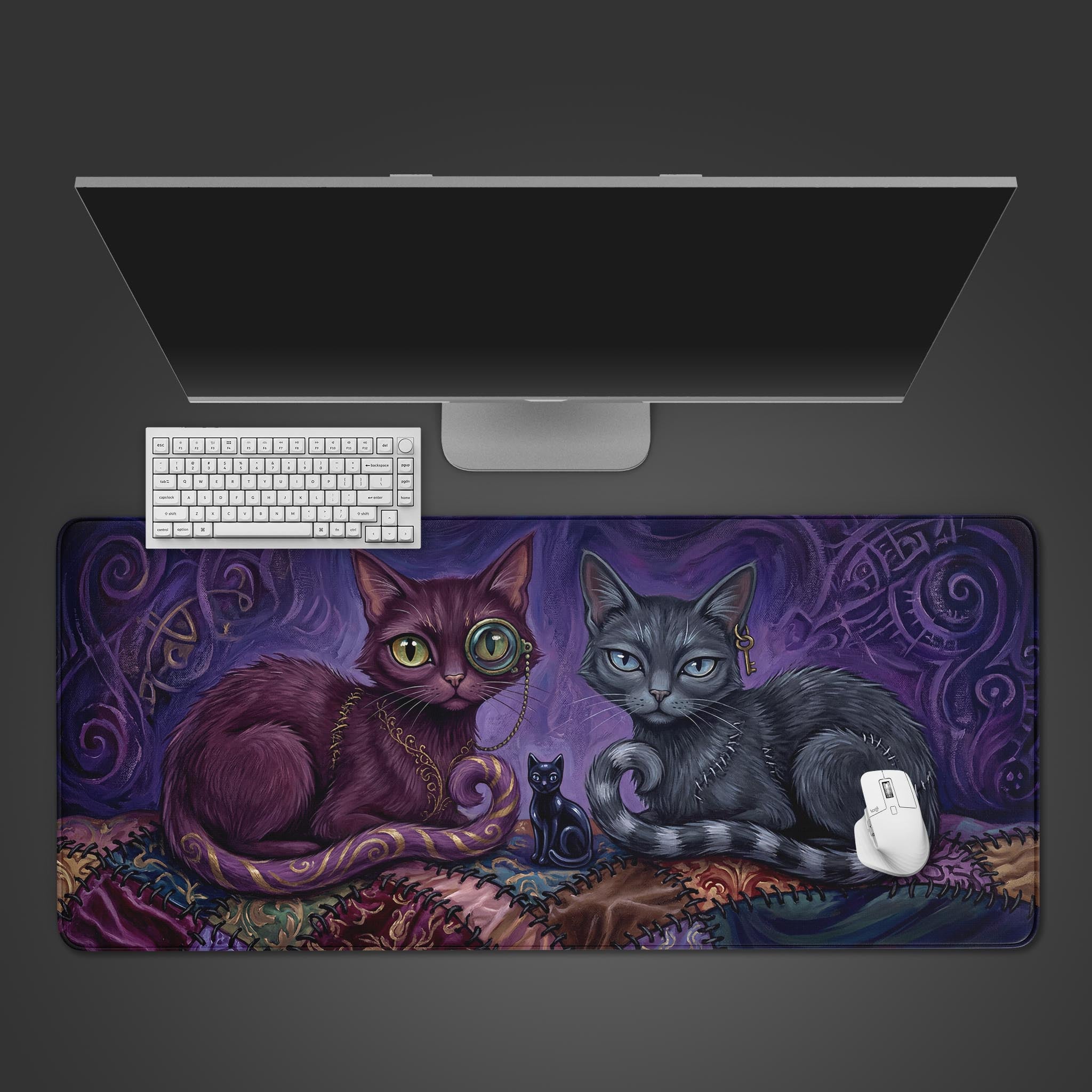 A top-down view of the Stitched Shadows gaming desk mat, featuring two mystical cats. A white keyboard and mouse are positioned on the mat below a large monitor. - Epic Design Pads & Mousepads